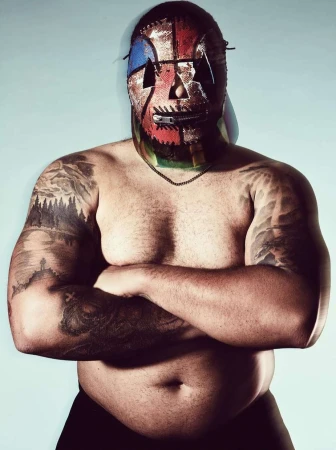 Man wearing a colorful stitched mask with a zipper mouth and heavy arm tattoos crossing his arms against a plain light background