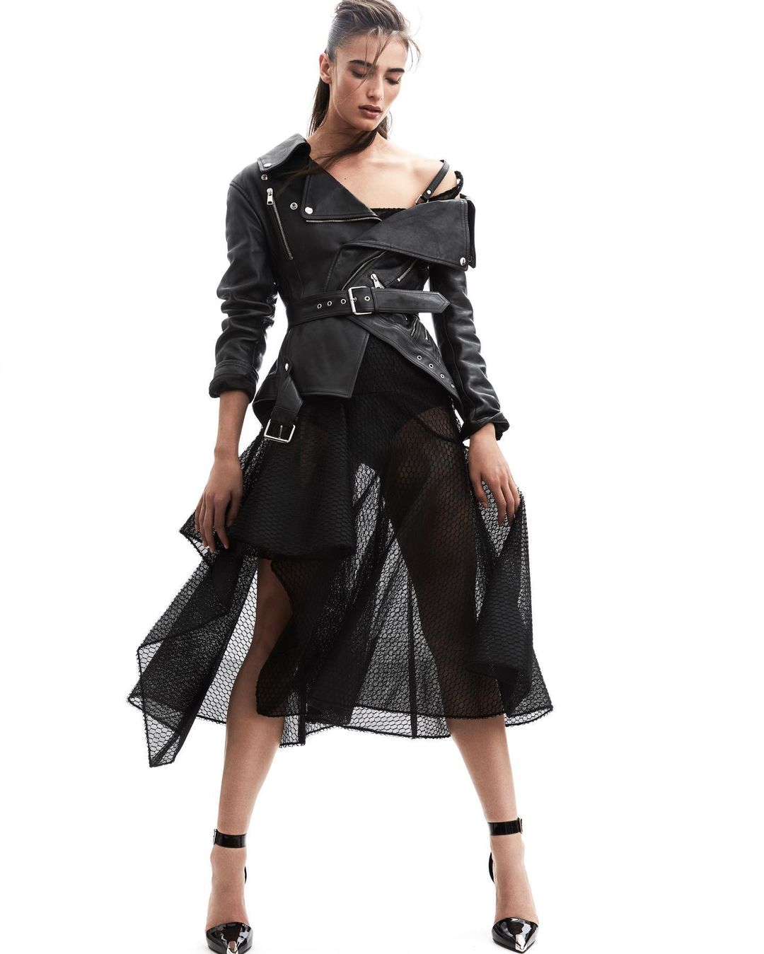 A woman posing in a black leather motorcycle jacket with a belted waist and a black mesh skirt on a white background.