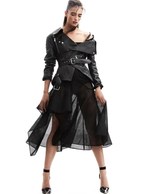 A woman posing in a black leather motorcycle jacket with a belted waist and a black mesh skirt on a white background.