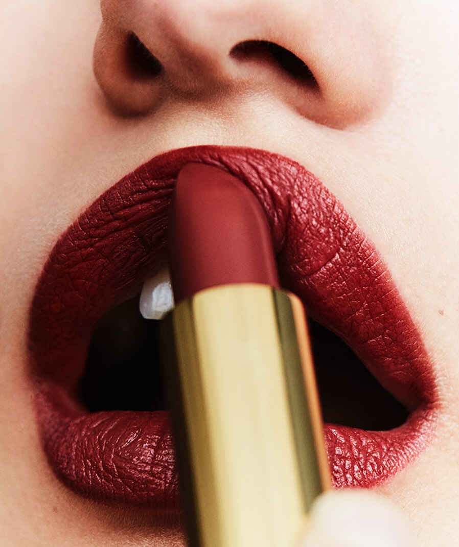 Close-up of a woman applying deep red lipstick from a gold tube, focusing on the lips, lipstick, and part of the nose.