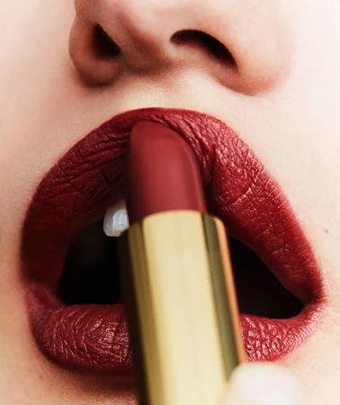 Close-up of a woman applying deep red lipstick from a gold tube, focusing on the lips, lipstick, and part of the nose.