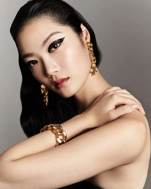 Glamour portrait of an Asian woman with bold eye makeup and golden jewelry, featuring a bracelet and dangling earrings on grey backdrop.