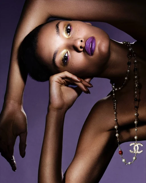 Glamorous close-up portrait of a dark-skinned woman with golden eyeshadow, purple lipstick, and a Chanel pearl necklace against a purple backdrop.