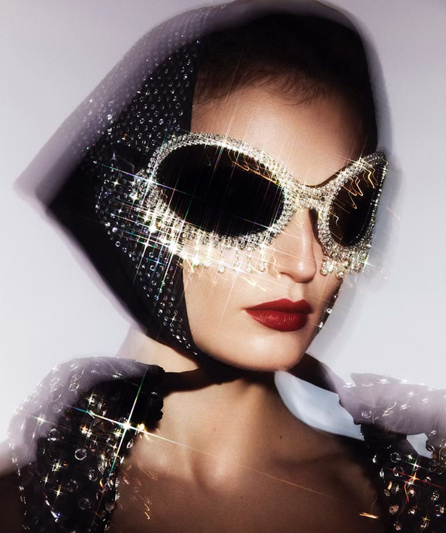 Glamorous woman with ruby red lips poses in a headscarf, embellished top, and embellished sunglasses against a plain backdrop. 
