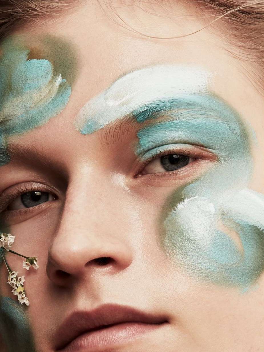 Close-up portrait of a person's face with light skin, adorned with white and teal paint strokes and a sprig of small white flowers.