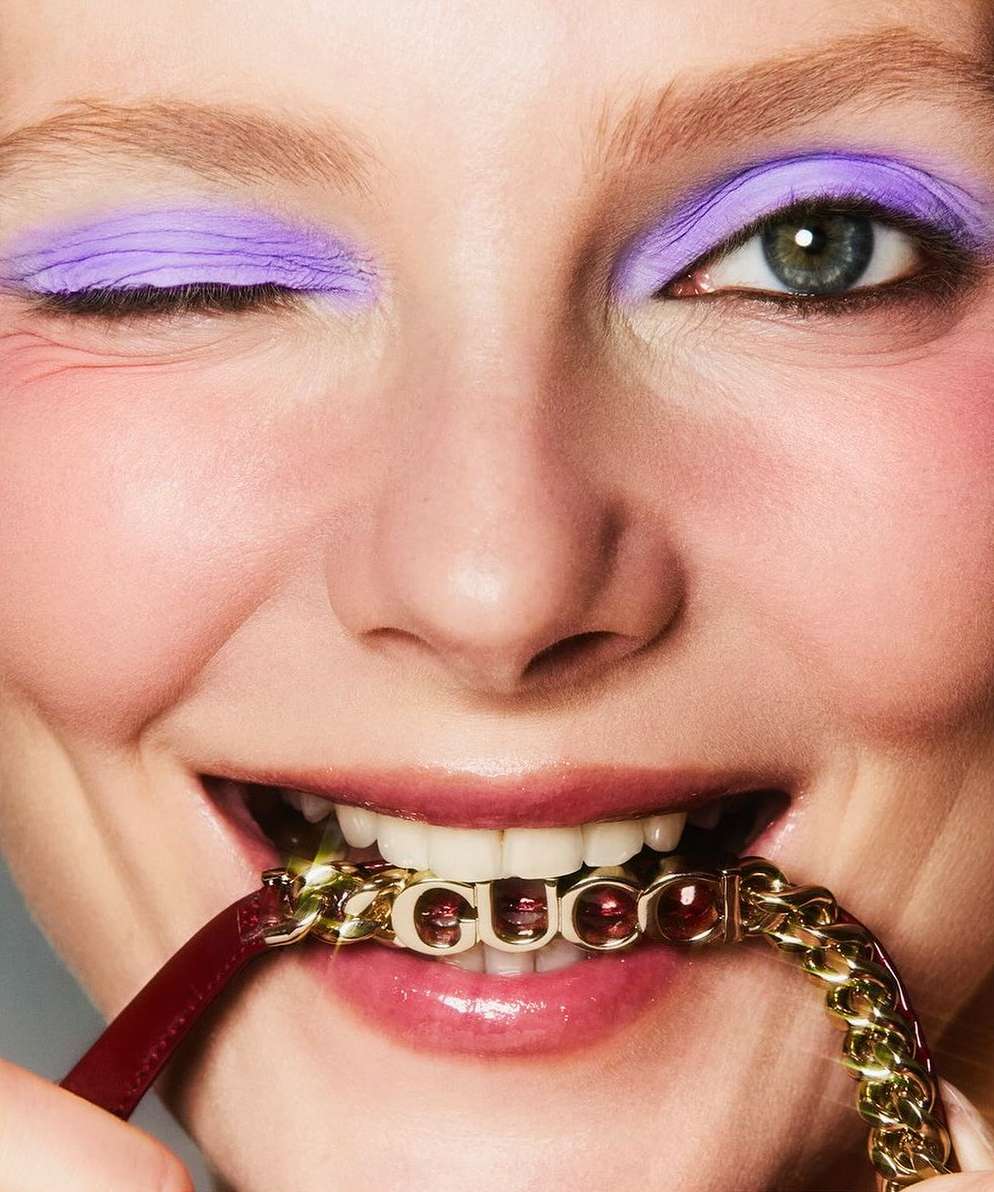 Close-up of a woman with lilac eyeshadow winking while biting down on a gold Gucci chain, showcasing beauty and high fashion.