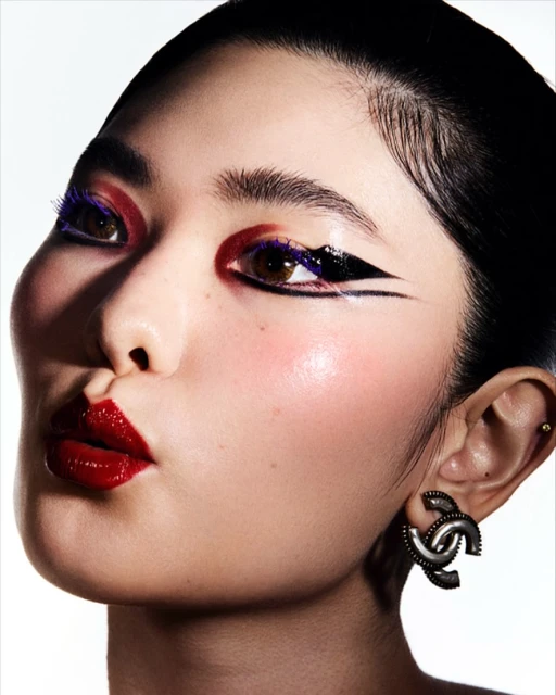 Close-up beauty shot of an Asian adult female model wearing colorful makeup and statement Chanel earrings against a white backdrop.