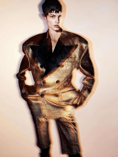 A model poses in a gold suit with exaggerated shoulders in a fashion forward image with a light background and layered visual effects.