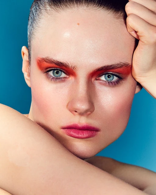 Close-up portrait of a young woman with dramatic red eye makeup and pink lipstick against a blue background, emphasizing beauty and fashion.