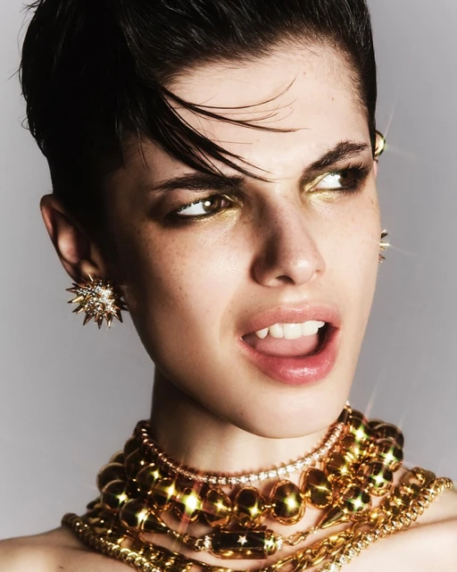 Close-up portrait of a young adult woman with short dark hair, heavy gold jewelry, and gold eye makeup, against a neutral background.