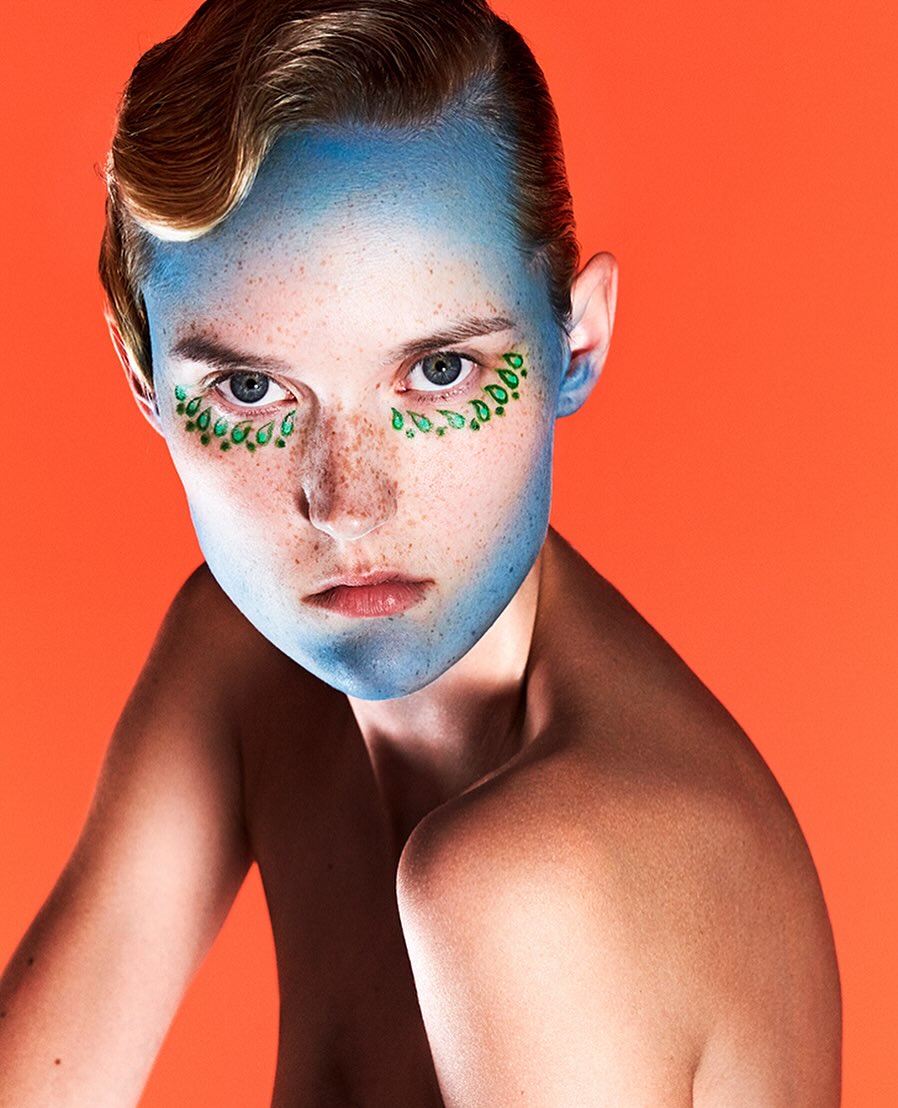 Portrait of a fair-skinned young adult with blue face paint and green leaf-shaped makeup designs around the eyes against an orange backdrop.