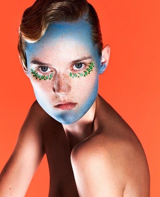 Portrait of a fair-skinned young adult with blue face paint and green leaf-shaped makeup designs around the eyes against an orange backdrop.
