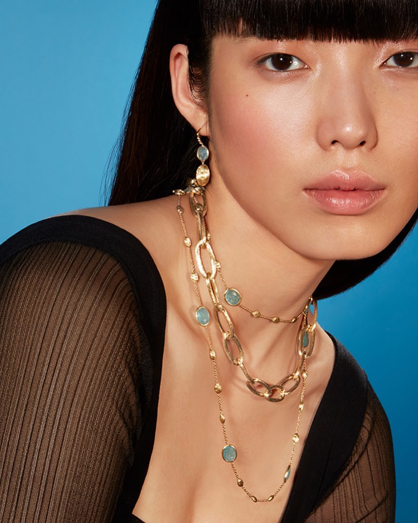 A close-up shot features an Asian woman with long dark hair wearing a sheer top and long gold chain necklaces against a blue background.