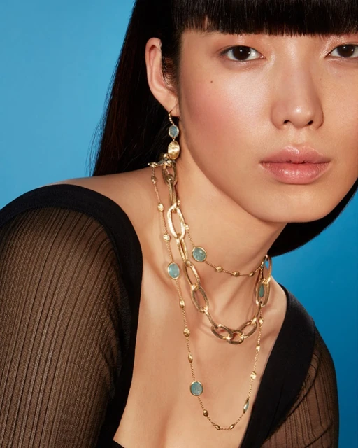 A close-up shot features an Asian woman with long dark hair wearing a sheer top and long gold chain necklaces against a blue background.