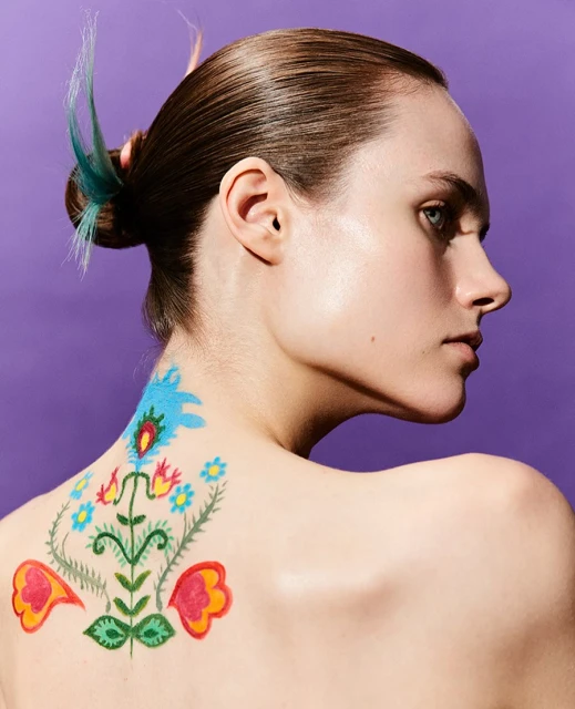 Woman with colorful flower body art on her back and blue highlight in hair poses against a purple background in a beauty shot.
