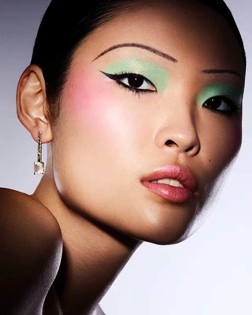 Close-up beauty shot of an Asian woman with vibrant mint green eyeshadow, exaggerated black eyeliner, and blush, accented by geometric earrings.