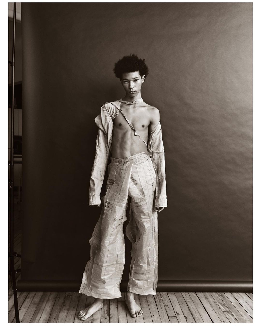 Person in newspaper fashion stands in photo studio. Young adult with afro, wearing deconstructed top and baggy pants. Grey backdrop.
