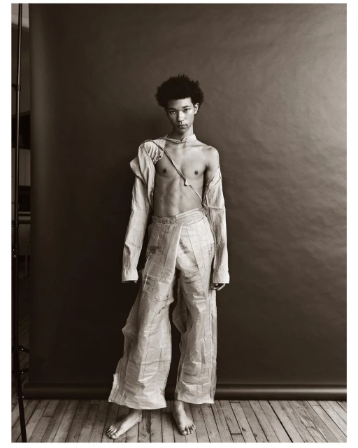 Person in newspaper fashion stands in photo studio. Young adult with afro, wearing deconstructed top and baggy pants. Grey backdrop.