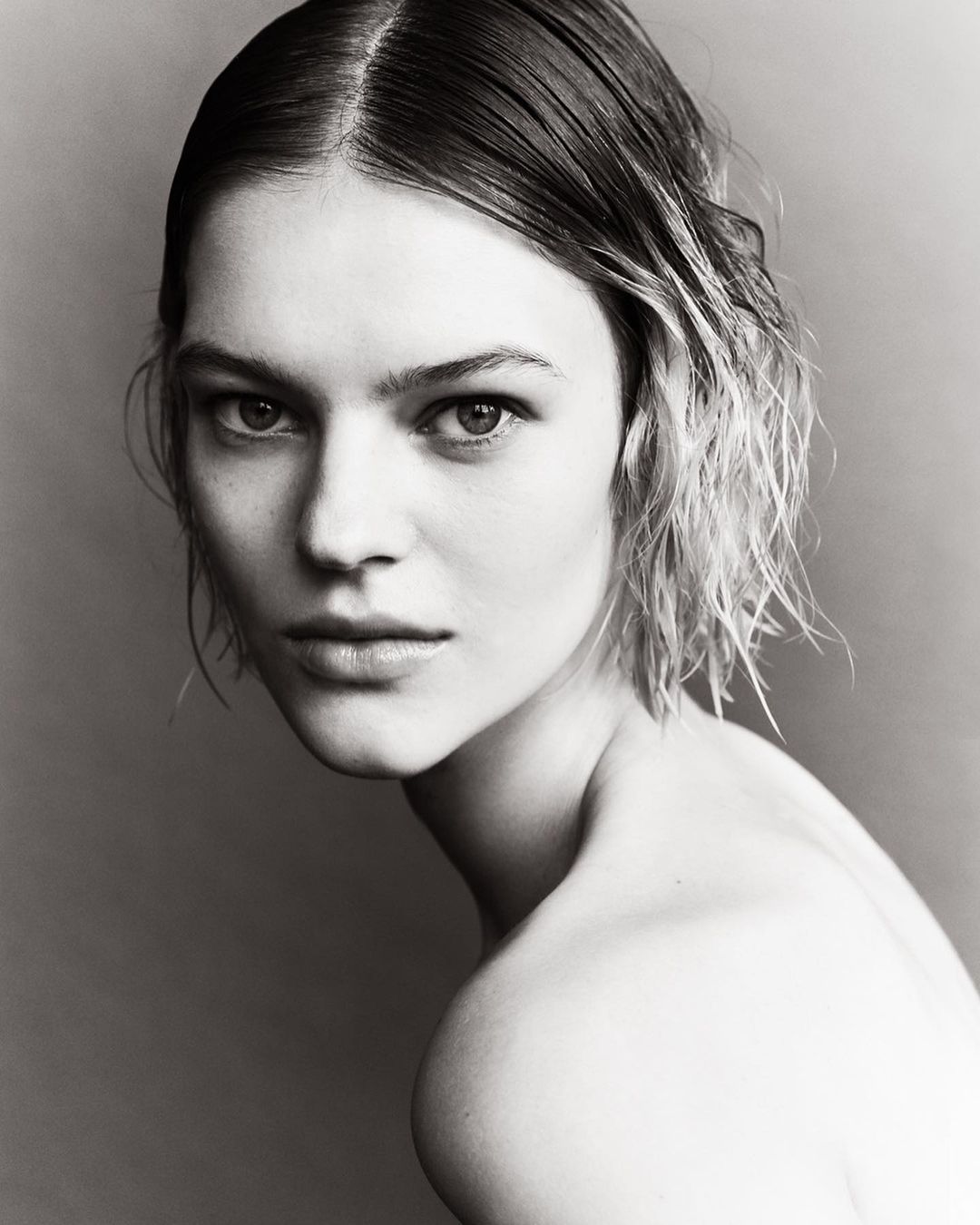 Black and white portrait of a woman with short, slicked-back hair, gazing directly at the camera with a neutral expression and bare shoulders.