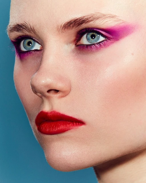 Close-up shot of a woman with fair skin, blue eyes, red lipstick, and purple eyeshadow against a blue background in a beauty editorial.