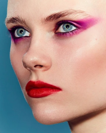 Close-up shot of a woman with fair skin, blue eyes, red lipstick, and purple eyeshadow against a blue background in a beauty editorial.
