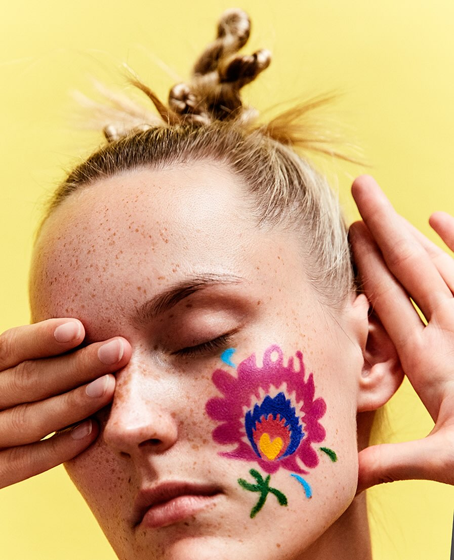 Close-up portrait of a woman with fair skin, freckles, and a stylized flower painted on her cheek, eyes closed, against a yellow backdrop.