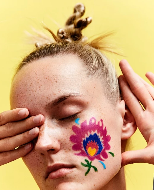 Close-up portrait of a woman with fair skin, freckles, and a stylized flower painted on her cheek, eyes closed, against a yellow backdrop.