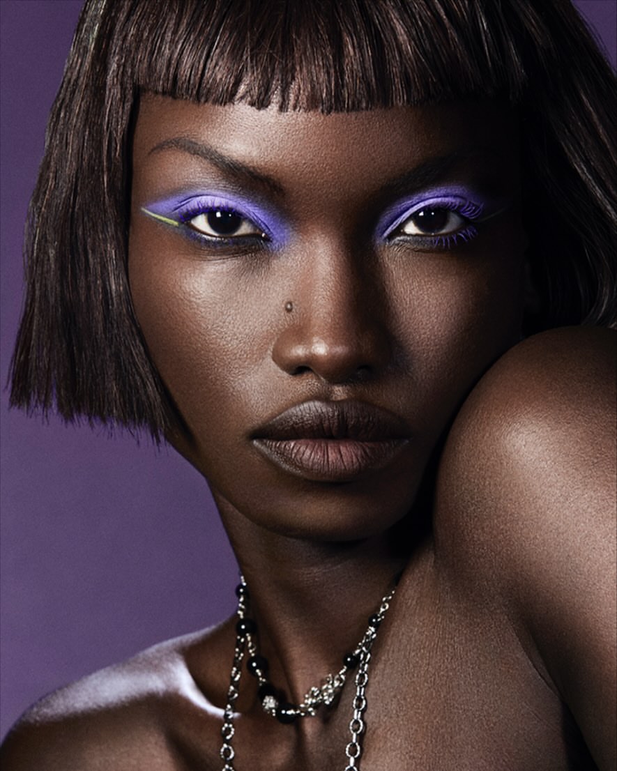 Close-up beauty shot of an African American woman with purple eyeshadow, bold eyeliner, and a beaded necklace against a purple backdrop.