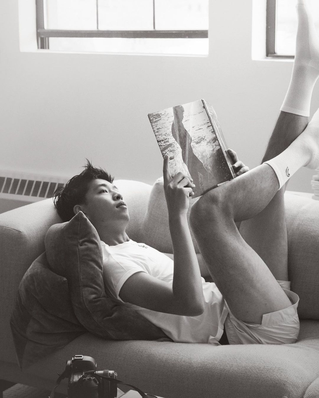An Asian male lies on a couch in white socks, reading a book with a landscape photo, with a camera on the couch.