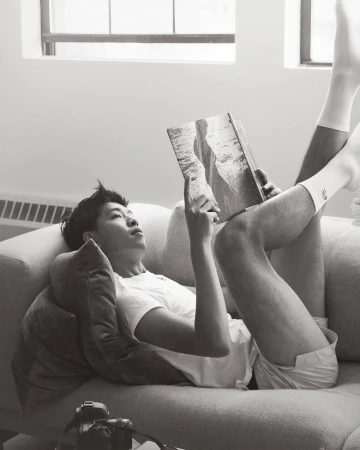 An Asian male lies on a couch in white socks, reading a book with a landscape photo, with a camera on the couch.