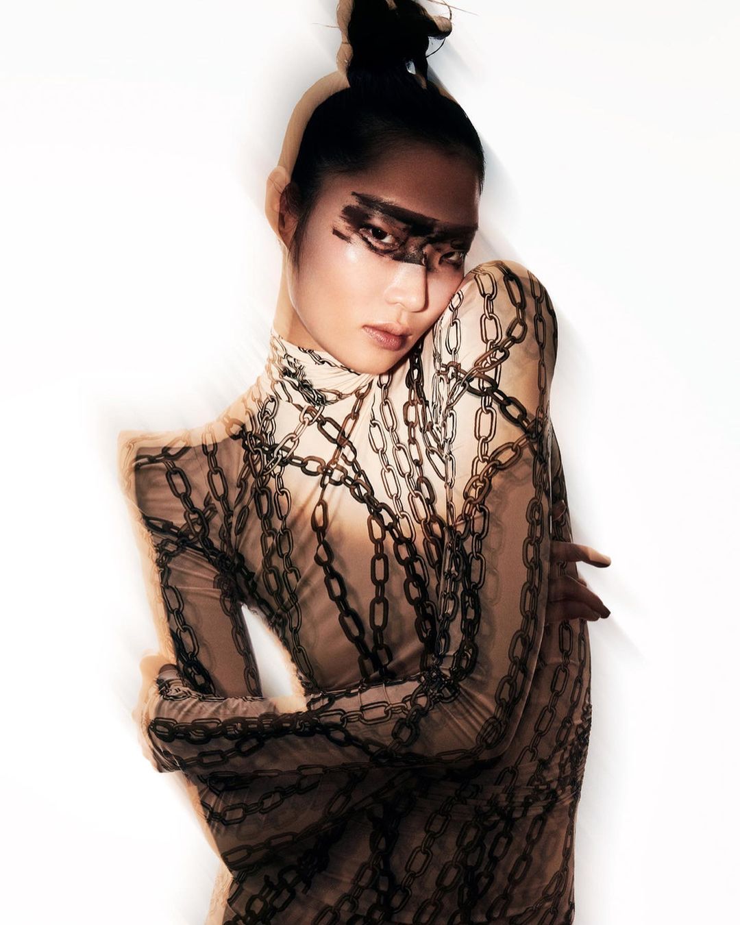 Elegant Asian woman with bold eye makeup wearing chain-print top against white background, posing with arms crossed in studio lighting.