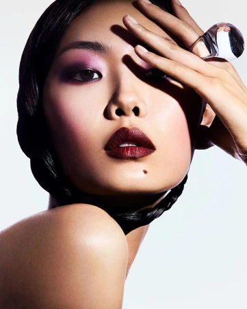 Close-up beauty portrait of an East Asian woman with dark hair, purple eyeshadow, dark red lipstick, and a large clear glass ring.