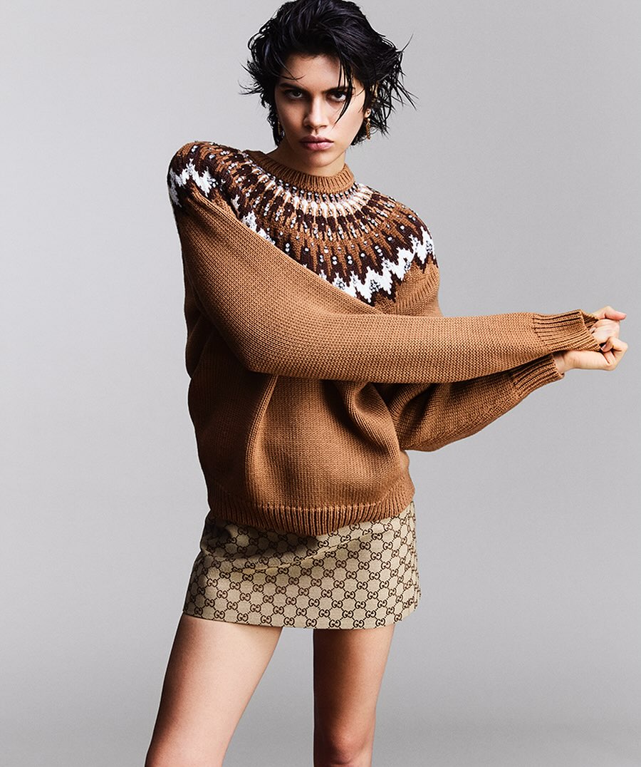 Fashion shot of a woman wearing a brown patterned sweater and logo skirt against a neutral studio backdrop, arms crossed and posing.