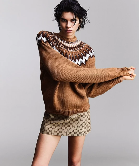Fashion shot of a woman wearing a brown patterned sweater and logo skirt against a neutral studio backdrop, arms crossed and posing.