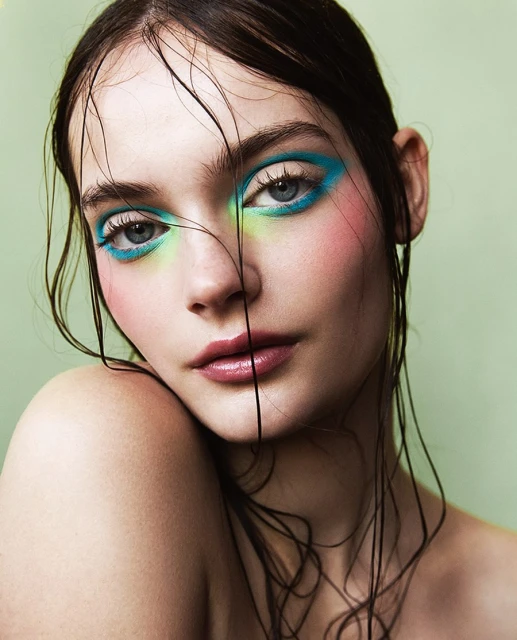 Close-up portrait of a young adult female model with vivid teal and green eye makeup against a light green background.