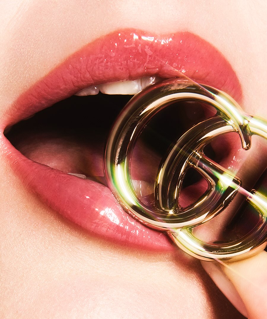 Close-up of a woman's glossy lips with a golden Gucci logo between her teeth, showcasing a beauty and fashion-forward aesthetic.