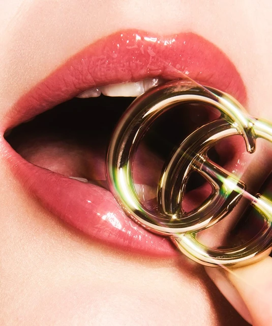 Close-up of a woman's glossy lips with a golden Gucci logo between her teeth, showcasing a beauty and fashion-forward aesthetic.
