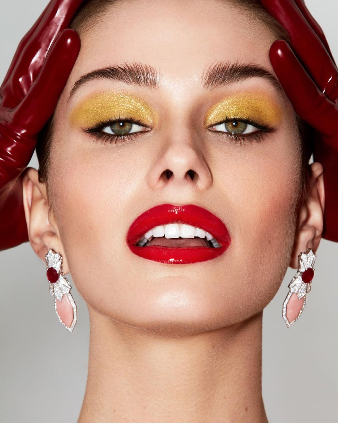Beauty shot of a woman with red lipstick and gold eyeshadow wearing sculptural earrings and red gloves against a neutral background.