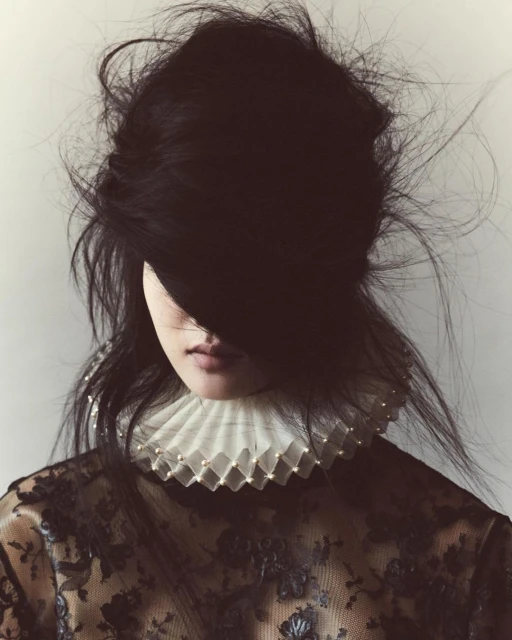 Portrait of a woman with black hair covering her face, wearing a lace top and ruffled pearl collar against a neutral background.