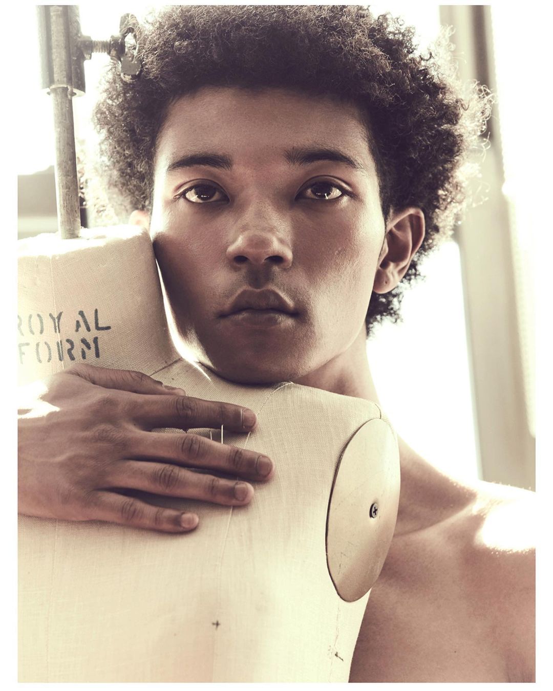 Portrait of a bare-chested man with dark curly hair embracing a tailor's dummy in a brightly lit studio setting, fashion or art photography.