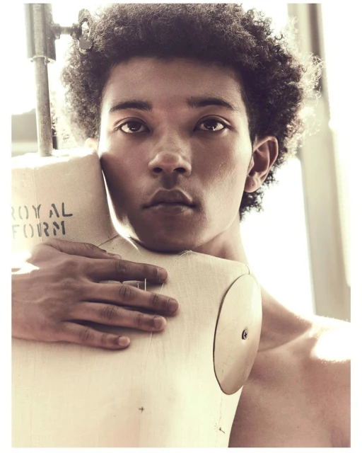 Portrait of a bare-chested man with dark curly hair embracing a tailor's dummy in a brightly lit studio setting, fashion or art photography.