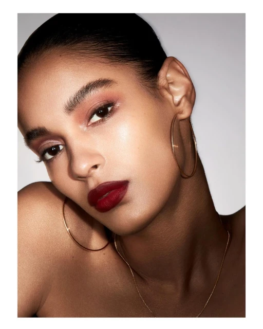Close-up portrait of a young, dark-haired woman with bronze skin, wearing large hoop earrings and dark red lipstick against a light grey backdrop.