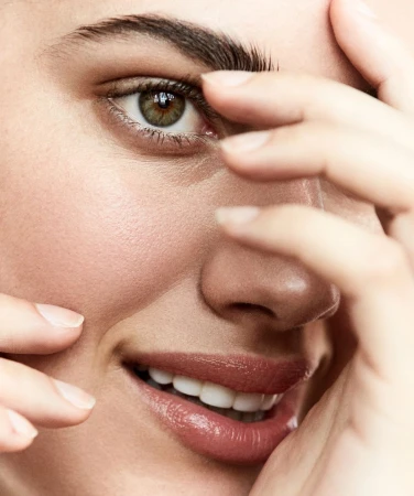 Close-up of a woman's face with fair skin, hazel eyes, and pink lips, partially obscured by her fingers. Beauty shot, smiling.