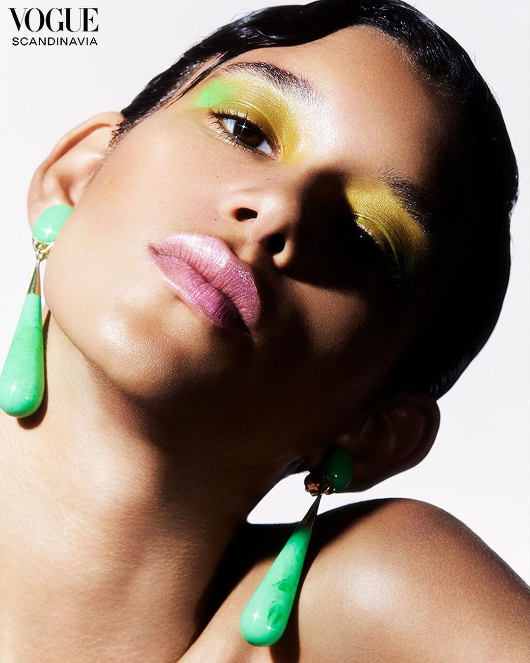 Close-up of a woman with glossy pink lips, wearing green eyeshadow, and hanging green earrings, against a bright white backdrop.