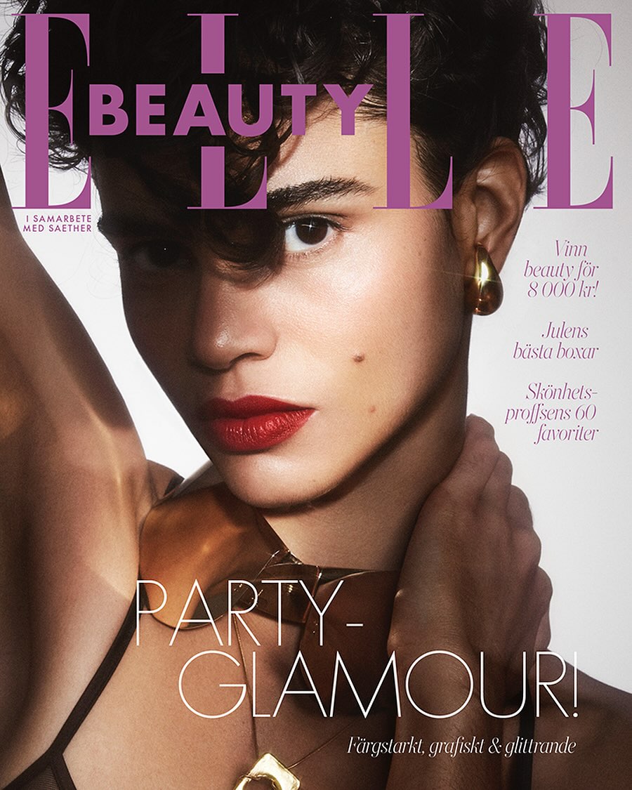 Elle Beauty magazine cover featuring a model with short, curly brown hair, red lipstick, gold earrings and necklace against a pink background.