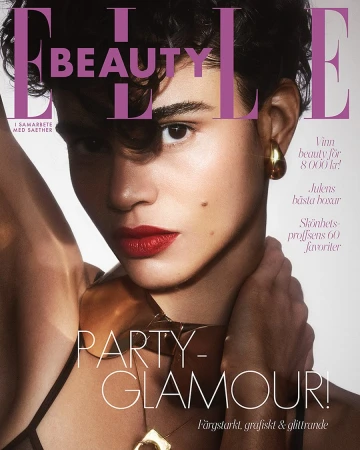 Elle Beauty magazine cover featuring a model with short, curly brown hair, red lipstick, gold earrings and necklace against a pink background.