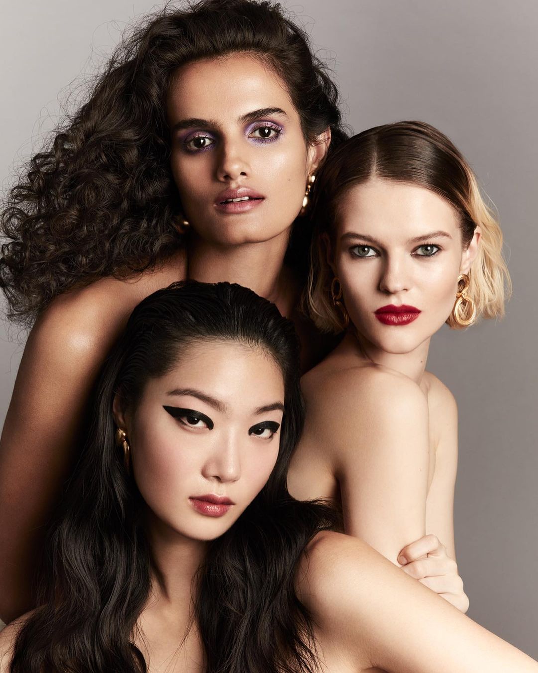Close-up studio portrait of three women of diverse ethnicities with bold makeup and gold earrings looking at the camera.