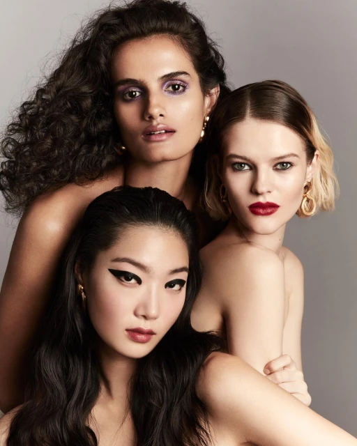 Close-up studio portrait of three women of diverse ethnicities with bold makeup and gold earrings looking at the camera.