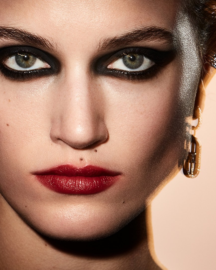Close-up portrait of a woman with dramatic black eye makeup and dark red lipstick, complemented by gold earrings. Beauty shot.