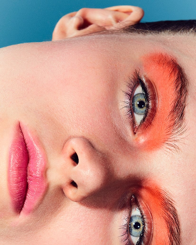 Close-up beauty shot of a model's face featuring vibrant coral eyeshadow and pink lips against a bright blue backdrop for a striking contrast.
